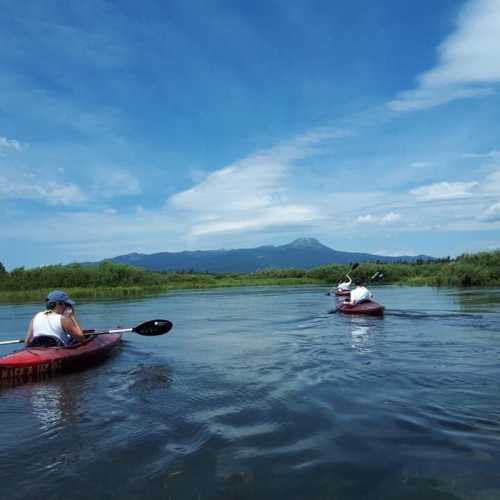 Two people kayak on a calm river surrounded by lush greenery and mountains under a clear blue sky.