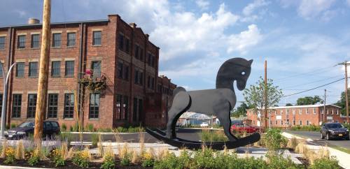 A large black rocking horse sculpture in a landscaped area with a brick building in the background and blue sky.