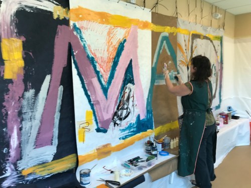 A person paints large abstract designs on paper panels in a creative workspace, surrounded by art supplies.