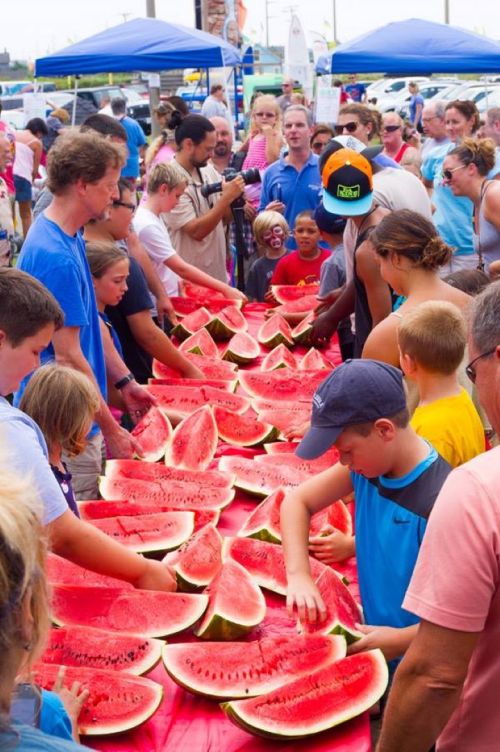 A large group of people at an outdoor event enjoying a watermelon feast, with many slices laid out on a table.