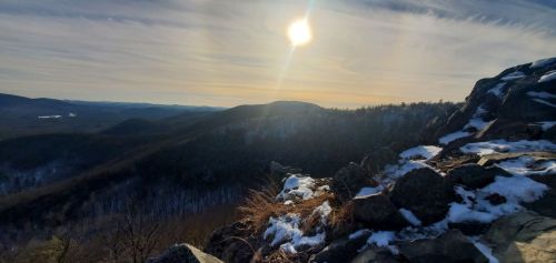 A scenic mountain view with snow-covered rocks, rolling hills, and a bright sun in a clear sky.