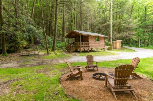 A cozy cabin in a forest clearing, surrounded by chairs and a fire pit, with a picnic table nearby.