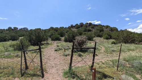 A dirt path leads through an open gate towards a rocky hill, surrounded by green shrubs and blue sky.