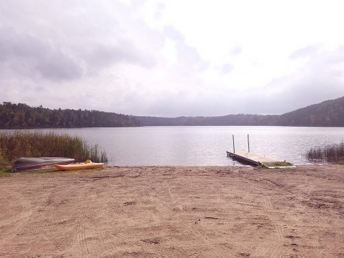 A serene lake view with a sandy shore, a wooden dock, and kayaks resting nearby under a cloudy sky.