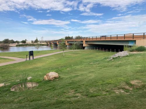 A grassy area with a bridge over a river, under a blue sky with scattered clouds. A path runs alongside the water.