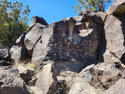 Ancient petroglyphs carved into a large rock face, surrounded by sparse vegetation and a clear blue sky.