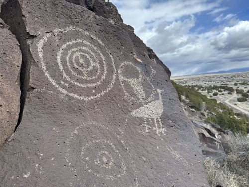Petroglyphs on a rock surface, featuring spiral designs and a figure resembling a bird or vessel, against a desert landscape.