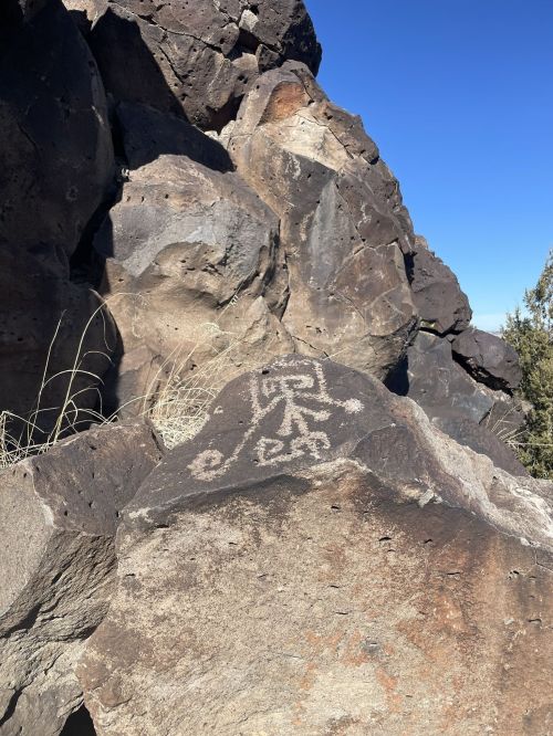 Petroglyph on a rock surface, featuring abstract designs, surrounded by rugged boulders and clear blue sky.