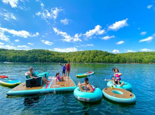 A group of children and adults enjoying a sunny day on inflatable rafts and a floating platform on a lake.
