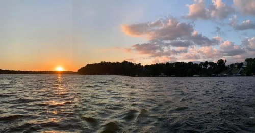A serene sunset over a lake, with gentle waves and a silhouette of trees and houses along the shore.