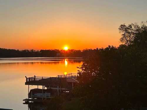 Sunset over a calm lake, reflecting orange and yellow hues, with a dock and trees in silhouette.