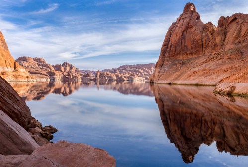 A serene landscape featuring red rock formations reflected in calm water under a blue sky with wispy clouds.