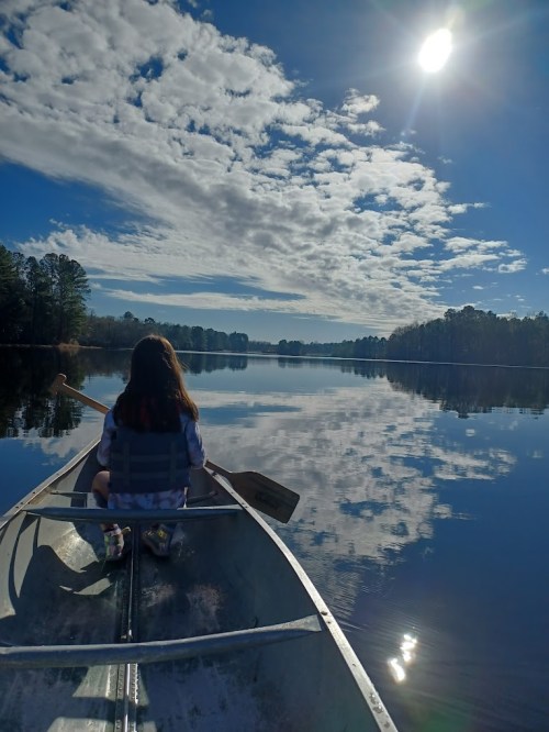 A person in a boat paddles on a calm lake, reflecting clouds and sunlight, surrounded by trees.