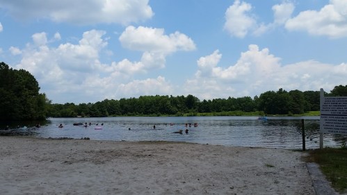 A sandy beach area by a calm lake with people swimming and a blue sky filled with fluffy clouds.