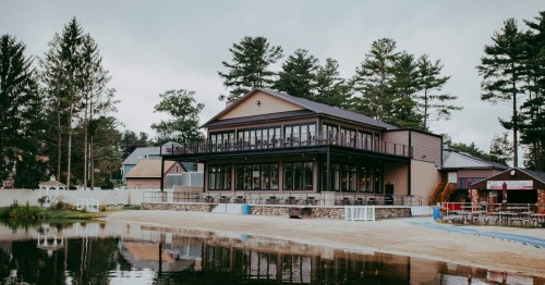 A modern lakeside building with large windows, surrounded by trees and a sandy beach area.