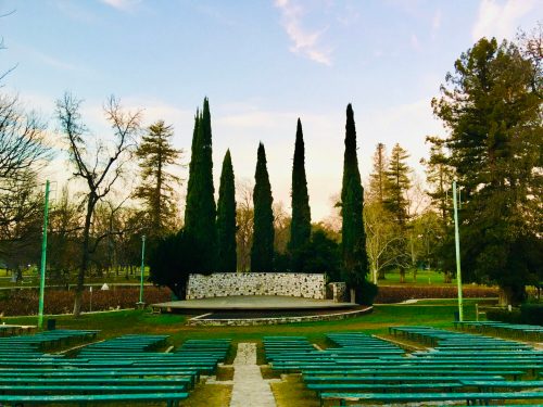 An outdoor amphitheater with green benches, a stone stage, and tall cypress trees against a colorful sky.