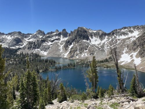 A scenic view of a mountain lake surrounded by rugged peaks and lush green trees under a clear blue sky.