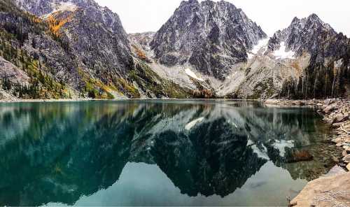 A serene mountain lake reflecting rugged peaks and autumn foliage under a cloudy sky.