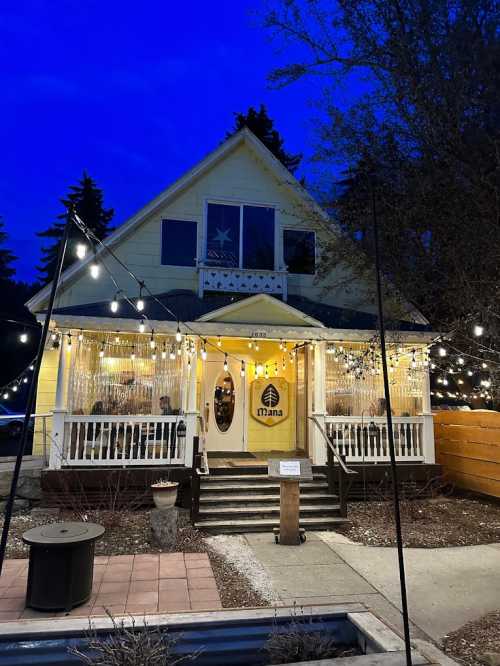 A charming yellow house with string lights, a welcoming entrance, and a sign reading "mana" against a twilight sky.