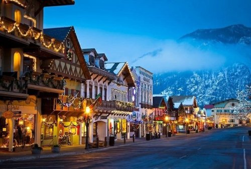 Charming alpine village street adorned with festive lights, surrounded by mountains under a twilight sky.