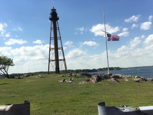 A tall lighthouse beside a grassy area with a flag at half-mast, overlooking a calm body of water under a blue sky.