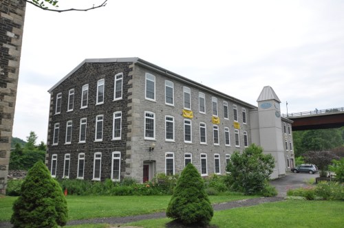 Historic stone building with large windows, surrounded by greenery, featuring a modern tower on one side.