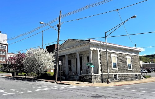 Historic building on a street corner, surrounded by blooming trees and power lines, under a clear blue sky.