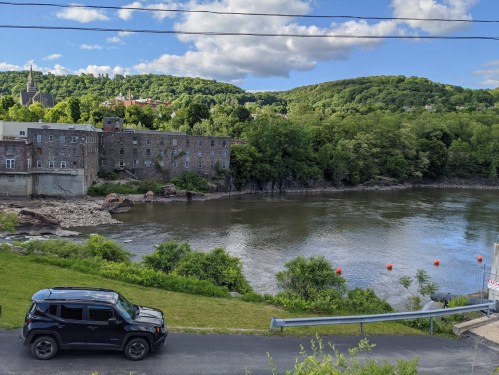 A black SUV parked by a river, with green hills and a stone building in the background under a blue sky with clouds.
