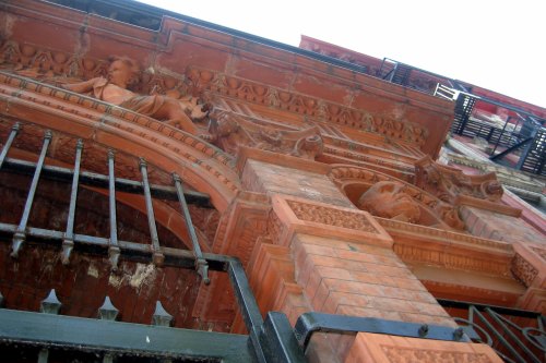 Close-up of ornate brickwork and sculptures on a historic building, viewed from below with a wrought iron gate.