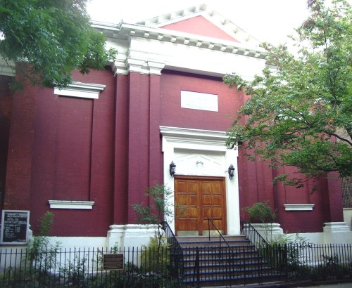 A red brick building with white columns and a large wooden door, surrounded by greenery and a wrought-iron fence.