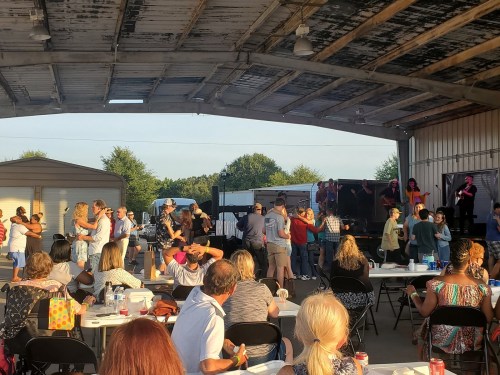 A lively outdoor event with people dancing and enjoying music under a large shelter, surrounded by tables and trees.