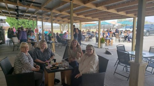 A group of four women sitting at a table outdoors, smiling, with a lively gathering in the background.
