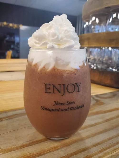 A glass of chocolate drink topped with whipped cream, featuring the text "ENJOY" and "Three Star Vineyard and Orchard."