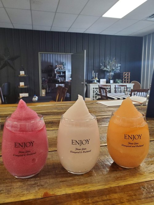 Three colorful frozen drinks in clear cups labeled "ENJOY," displayed on a wooden table in a cozy interior setting.