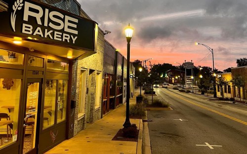 A bakery storefront at dusk, with a colorful sunset and a quiet street lined with trees and streetlights.