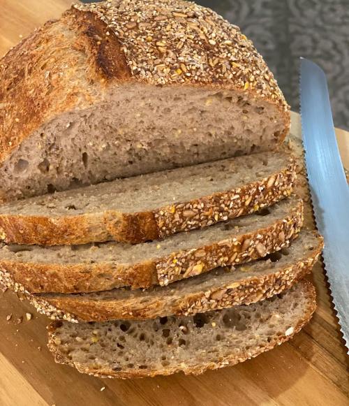Sliced whole grain bread with a crusty top, sprinkled with seeds, on a wooden cutting board next to a serrated knife.