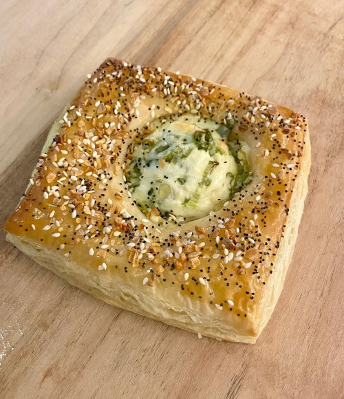 A flaky pastry square topped with sesame seeds and poppy seeds, featuring a creamy filling in the center.