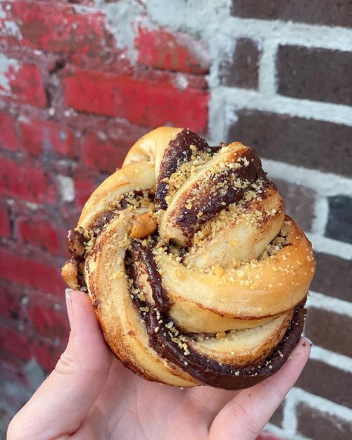 A hand holding a twisted pastry with chocolate filling and a crumbly topping against a brick wall background.