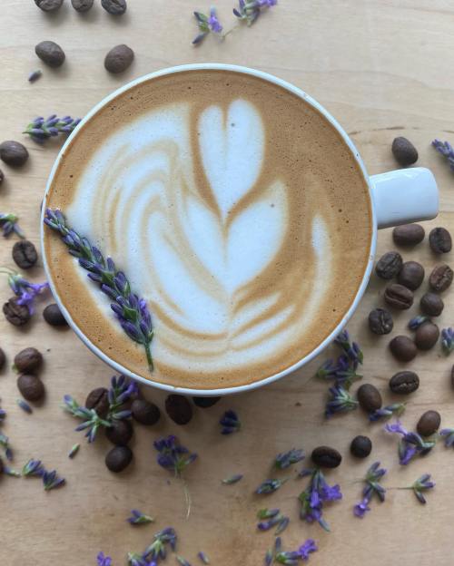A latte with a floral design, topped with lavender, surrounded by coffee beans and lavender petals on a wooden surface.