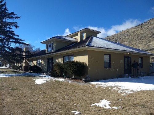 A yellow house with a sloped roof, surrounded by grass and patches of snow, set against a blue sky and hillside.