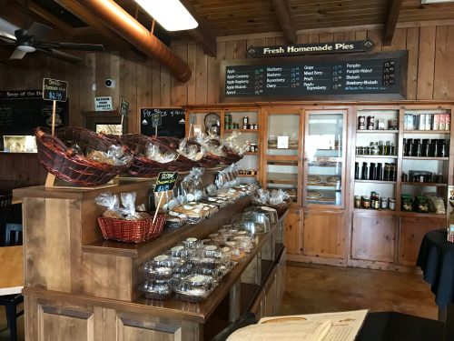 A cozy bakery display with baskets of baked goods, jars of preserves, and a glass case filled with pies. Warm wooden decor.