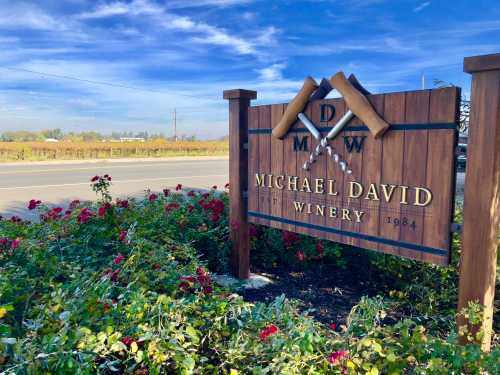 Sign for Michael David Winery, featuring crossed wine barrels, surrounded by flowers and vineyards under a blue sky.