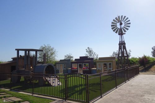 A playground featuring a wooden train structure, small houses, and a windmill, surrounded by green grass and a fence.