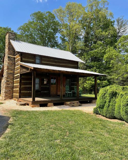 A rustic log cabin with a metal roof, surrounded by greenery and trees, featuring a porch and stone chimney.