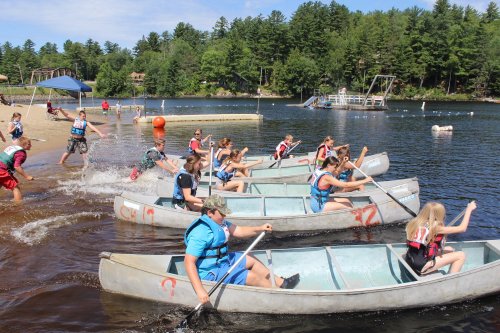 A group of campers paddles canoes on a lake, with trees and a sandy beach in the background.
