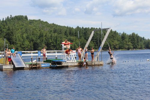 A lively lakeside scene with people swimming, diving, and enjoying a sunny day at a dock surrounded by trees.