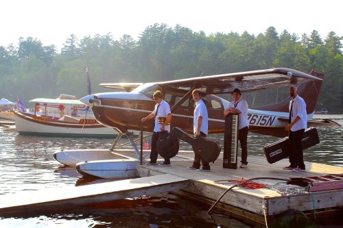 Four musicians in white shirts carry instruments off a seaplane onto a dock by a lake, surrounded by boats and trees.