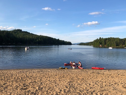 A serene lake scene with sandy beach, kayaks, and people enjoying the water surrounded by lush green hills.