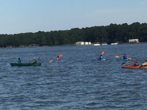 A group of kayakers paddles on a calm lake surrounded by trees and distant buildings.
