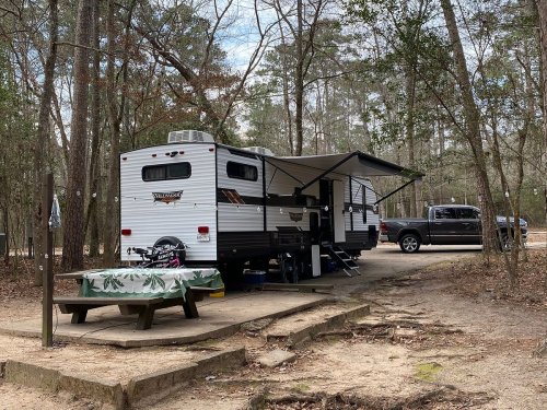 A camper trailer parked in a wooded campsite, with a picnic table and a pickup truck nearby.